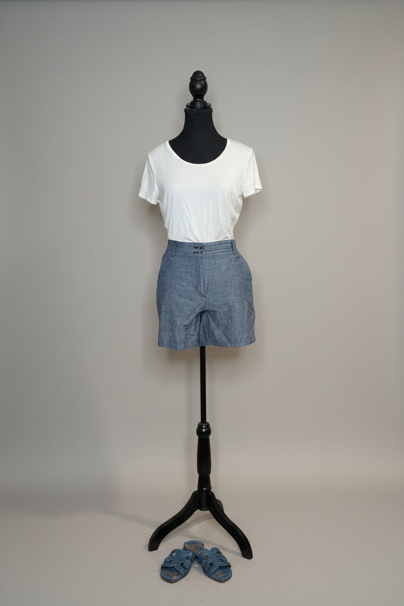 Mannequin wearing a white t-shirt and blue shorts against a gray wall with a vase of flowers on a table.