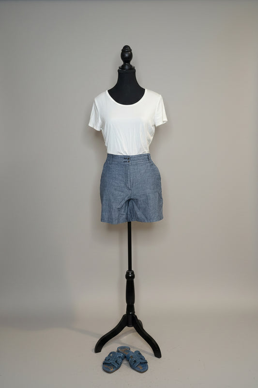 Mannequin wearing a white t-shirt and blue shorts against a gray wall with a vase of flowers on a table.